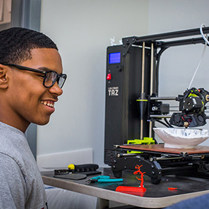 Student smiling while operating a 3D printer, creating colorful printed parts in a classroom.