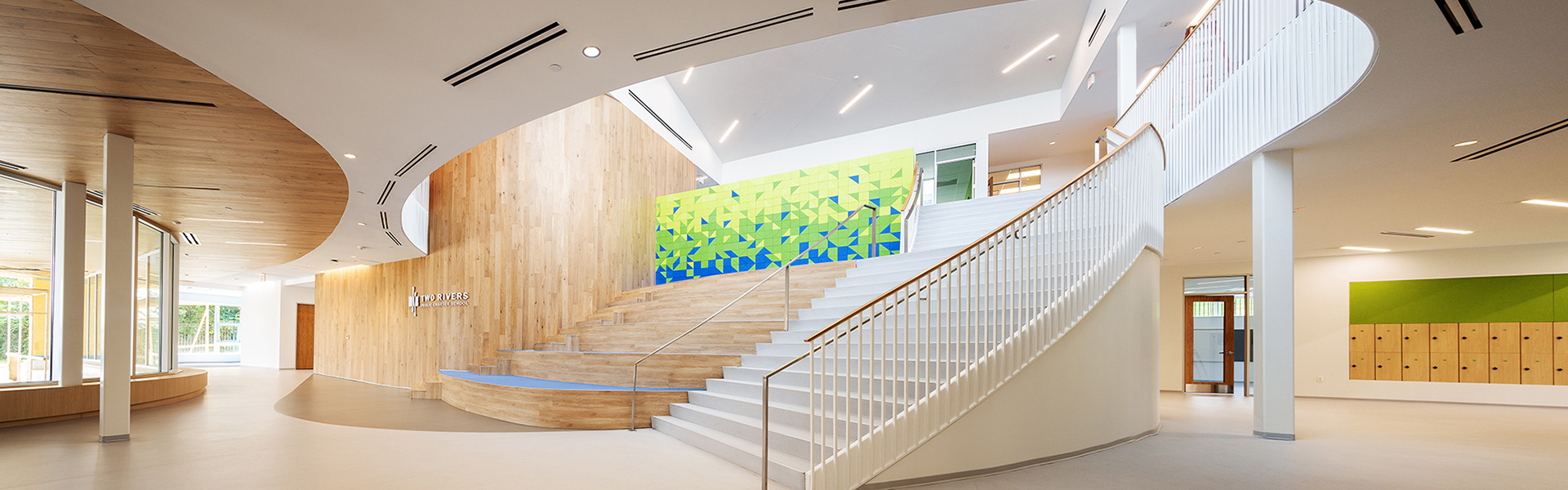 Spacious modern school atrium with curved staircase, wood accents, and colorful geometric wall art.