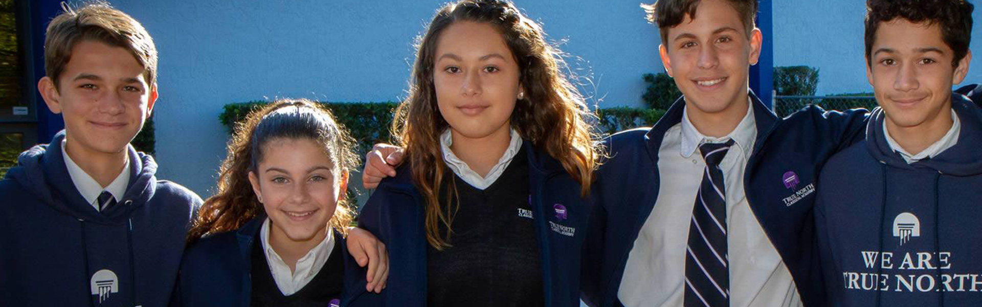 Five students in school uniforms smiling outdoors, wearing sweatshirts reading “TRUE NORTH.”