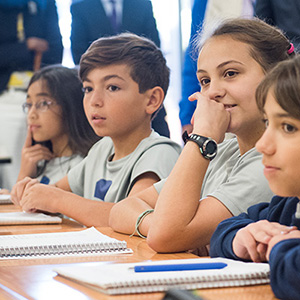 Four students sit at a table with notebooks, listening attentively in class.