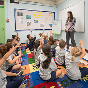 Teacher leads students on an alphabet rug as children raise their hands enthusiastically.