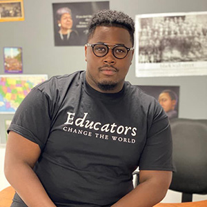 A man wearing glasses and a black "Educators Change the World" t-shirt sits in an office, surrounded by educational and inspirational posters.