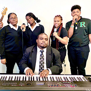 A group of four young adults and one man pose with musical instruments. The man sits at a keyboard, while the others hold microphones, conveying a musical theme.