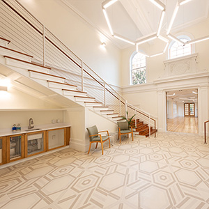 Spacious, modern lobby with geometric tile floor, a minimalistic staircase, two chairs, and a kitchen area. Soft lighting creates a welcoming atmosphere.