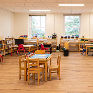 A bright, spacious classroom with wooden floors and tables. Shelves hold educational materials. Red and blue chairs add color. Large windows offer natural light.