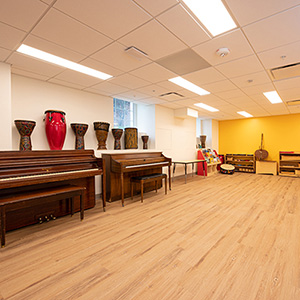 Spacious music room with two pianos, a row of drums, and various percussion instruments against a vibrant yellow wall, under bright ceiling lights.