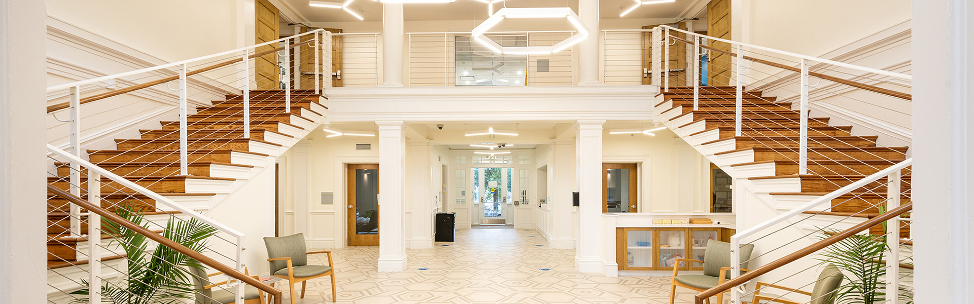 Bright, modern lobby with dual wooden staircases leading up. Geometric light fixtures hang from the ceiling. Green chairs and plants add a welcoming touch.