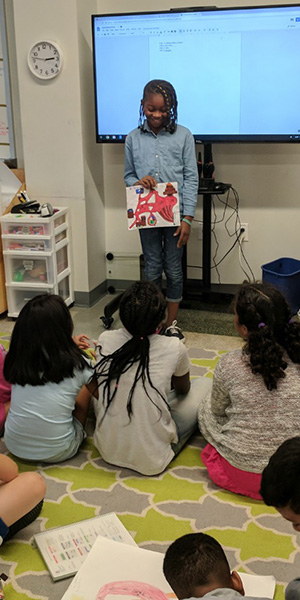 A child presents artwork to seated classmates in a classroom. The child stands in front of a large screen while peers sit attentively, creating an engaged atmosphere.