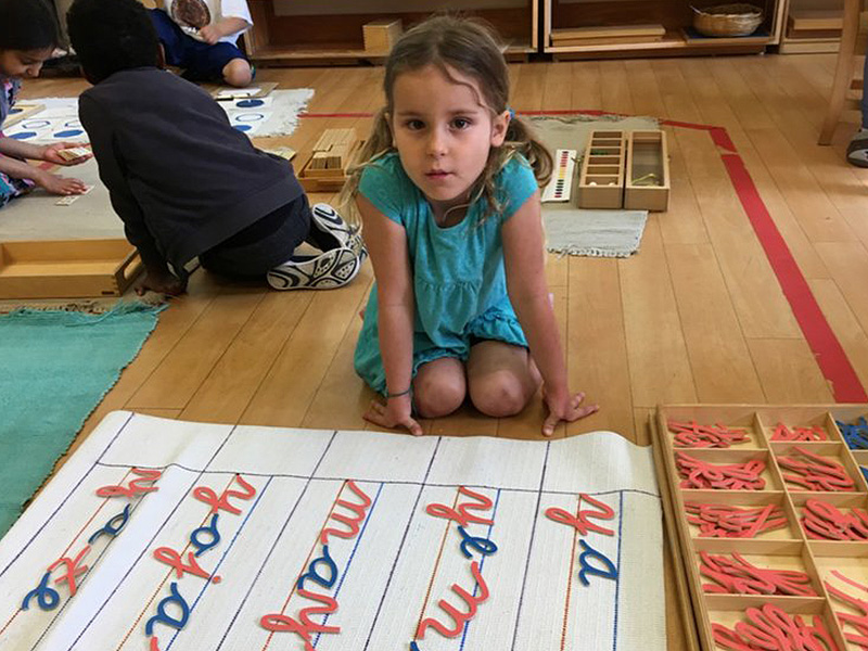A young girl in a turquoise dress sits on a classroom floor, surrounded by educational materials. Nearby, children engage in activities. The scene conveys focus and learning.