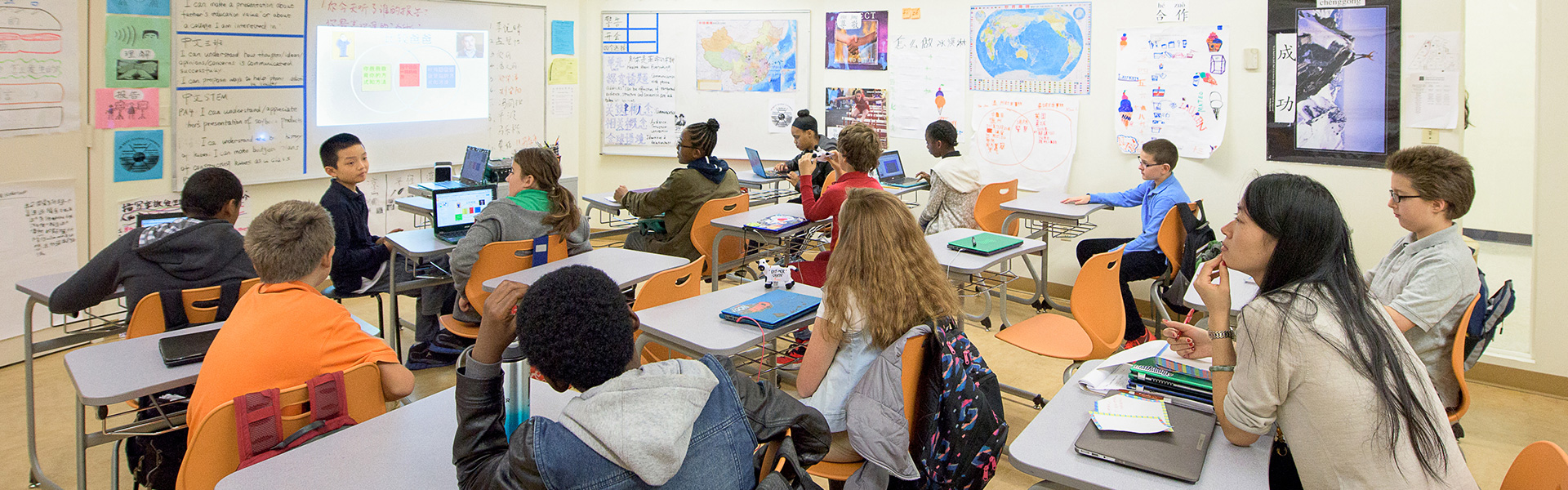 A classroom with students seated at desks, engaged and attentive to a boy presenting at the front. The walls are decorated with maps and posters.