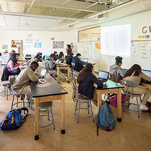A classroom with diverse students seated at desks, using laptops. A teacher stands at the front, projecting notes on a whiteboard. The atmosphere is focused.