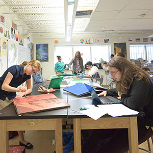 A classroom with students engaged in various activities at tables; one draws, another uses a laptop. The room is bright and filled with artwork.