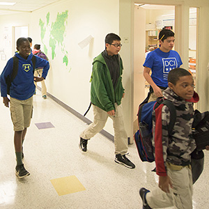 Four students walk down a school hallway. The walls are white with a green world map. They wear casual clothes and carry backpacks, conveying a lively school atmosphere.