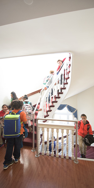 Children with backpacks ascend a well-lit, curved staircase with white railings. The mood is bustling as they prepare for school, some pausing to chat.