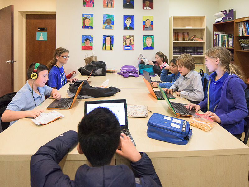 Students sit around a table with laptops in a classroom, focused and engaged. Colorful portraits adorn the wall, and bookshelves are in the background.