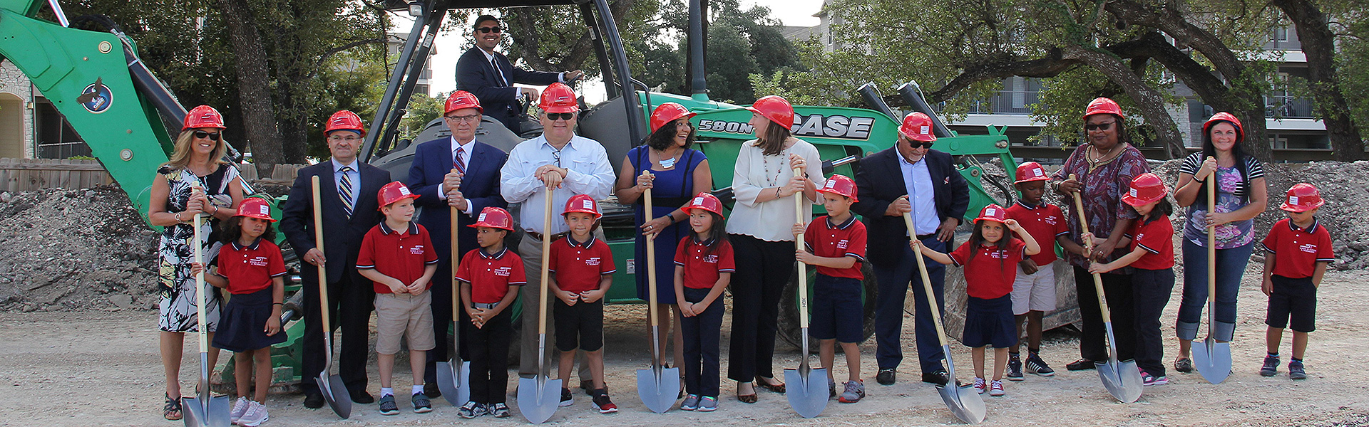 A diverse group of adults and children, all wearing red hard hats, stand in front of a green excavator on a construction site. They are holding shovels and smiling, suggesting a ceremonial groundbreaking event.
