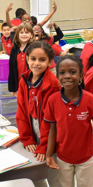A group of cheerful children in red uniforms gather in a classroom, smiling at the camera. The atmosphere is energetic and lively, with colorful folders around.