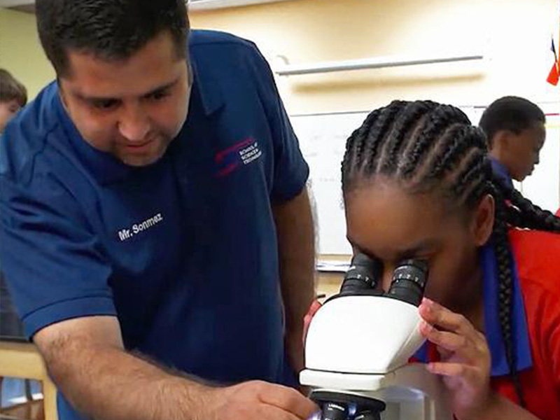 A teacher assists a student using a microscope in a classroom. The girl, focused and curious, peers into the lens. The scene conveys learning and guidance.