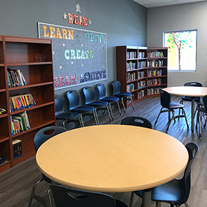Cozy library room with round tables, blue chairs, and wooden bookshelves filled with books. A bulletin board displays "READ, LEARN, CREATE."