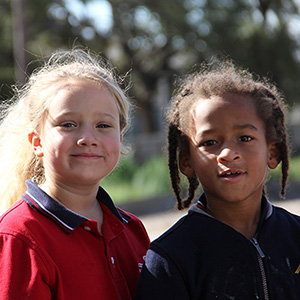 Two children stand outdoors, smiling softly. One wears a red shirt, the other a dark jacket. Sunlight filters through trees, creating a warm, cheerful atmosphere.