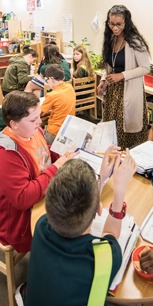 A lively classroom scene with students discussing at a round table, books open. A teacher stands nearby, smiling warmly. The setting feels collaborative.