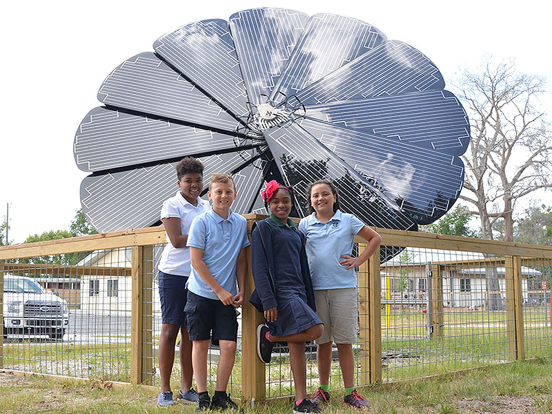 Four smiling children in school uniforms stand in front of a large solar panel shaped like a flower. The setting is outdoors, conveying a sense of innovation and sustainability.