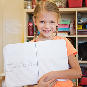 A young girl with braided hair smiles while holding an open notebook in a classroom. The room has shelves filled with colorful supplies, creating a cheerful atmosphere.