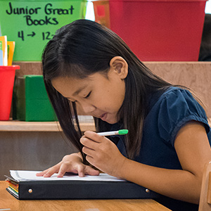 A young girl with long hair is focused on writing in a notebook at her desk. Behind her are colorful book bins. The setting is a classroom.