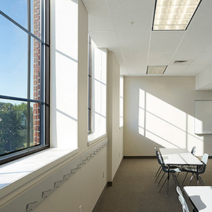 Sunlit classroom with large windows casting shadows on white walls. Desks and chairs are arranged neatly. The room feels bright and spacious.