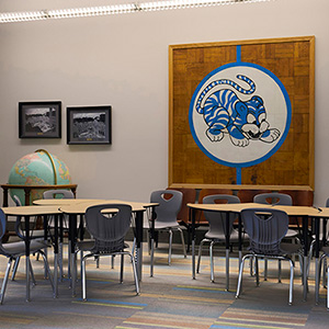 Classroom with circular tables and gray chairs arranged neatly. A large tiger mural adorns the wall, and a globe sits atop a cabinet. Walls are light-colored with framed photos. Quiet and organized ambiance.