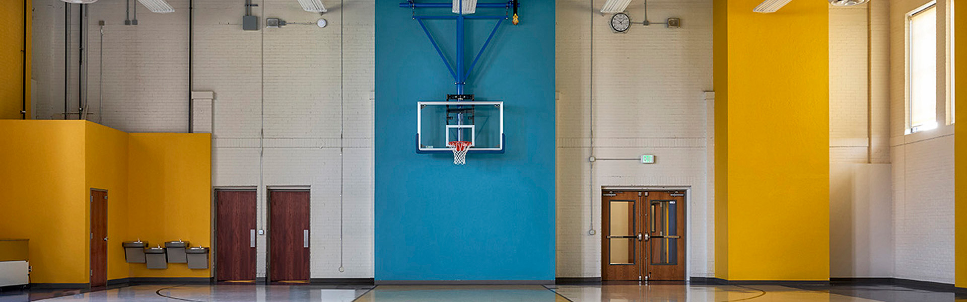 Spacious gym with a vibrant yellow and blue wall, featuring a basketball hoop and polished floor. Doors and water fountains on the left. Bright, open atmosphere.