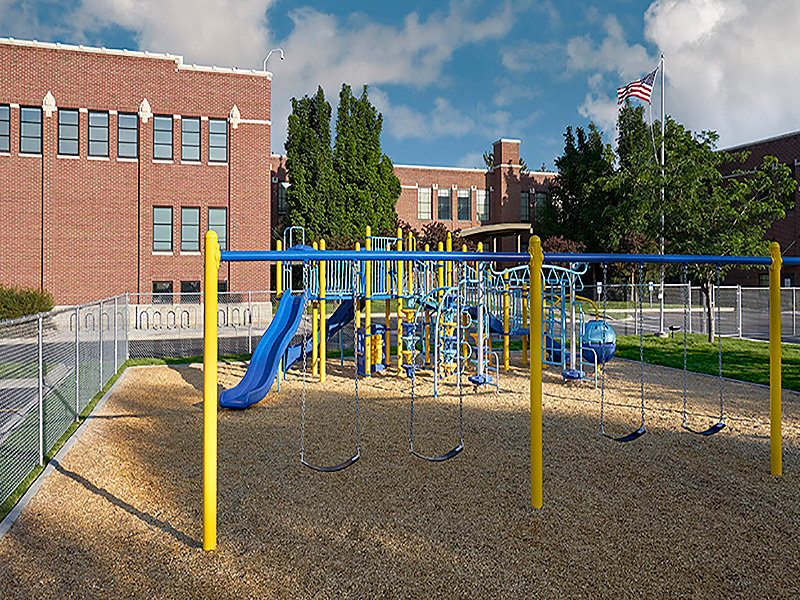 A school playground with colorful play structures, including swings and a slide, sits beside a brick building on a sunny day with an American flag.
