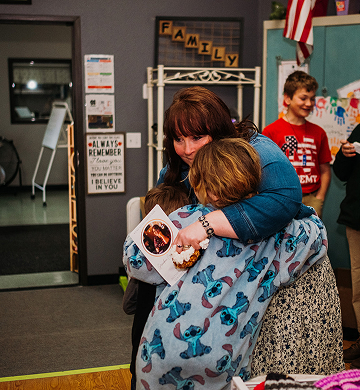 A woman in a denim jacket and a child wrapped in a blanket with Stitch print hug in a cozy room. They hold a cupcake and card. The mood is warm and affectionate.