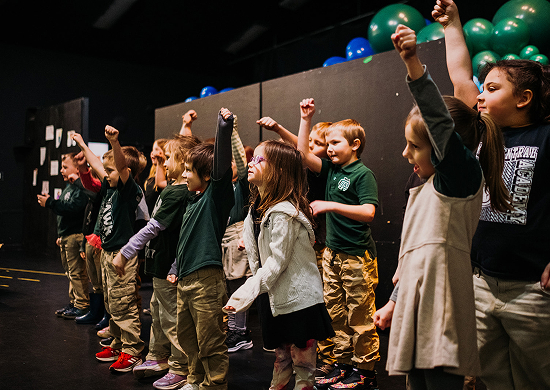 A group of children stand on a stage with colorful balloons behind them, their fists raised in unison. They appear enthusiastic and joyful.