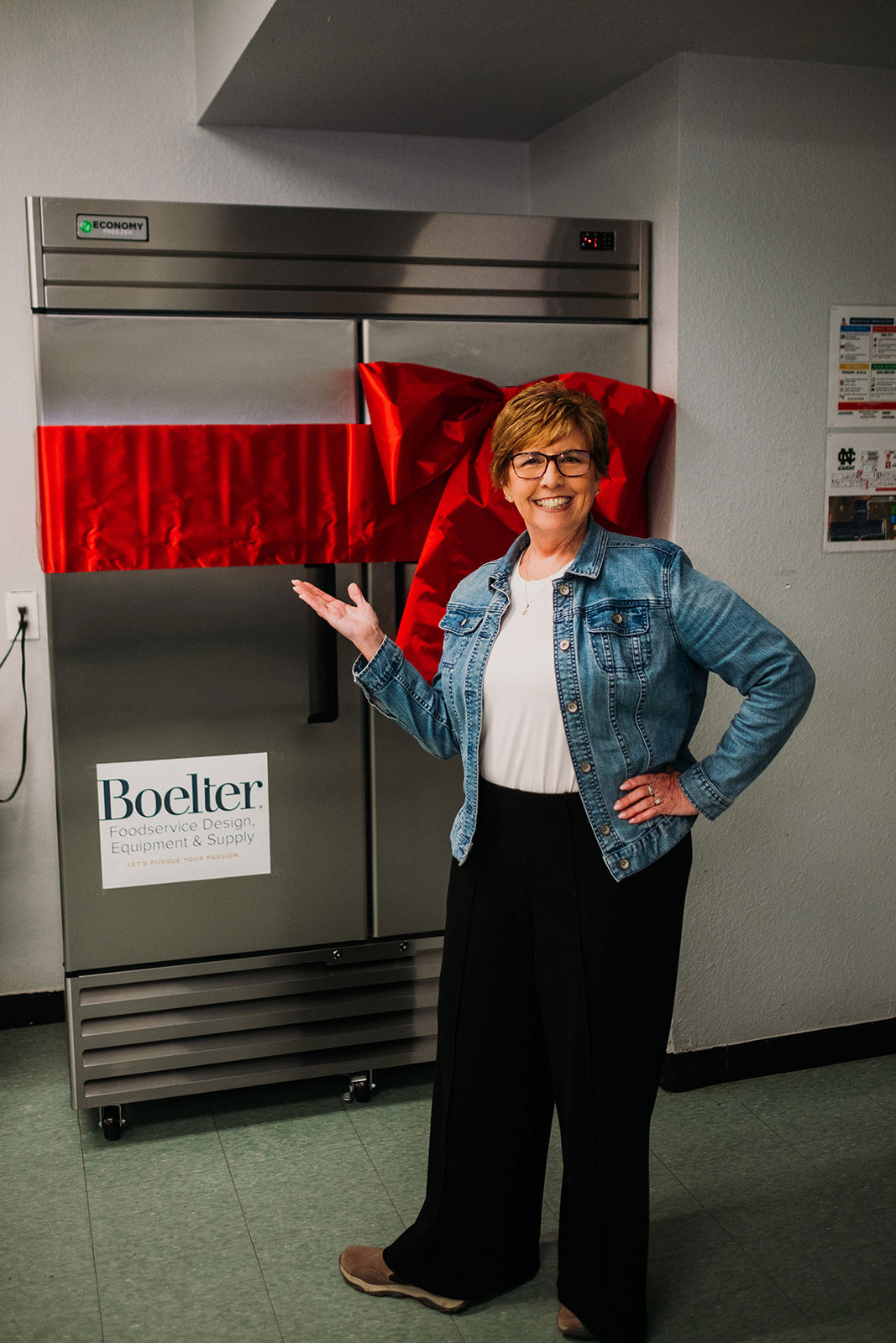 A woman in a denim jacket smiles, gesturing towards a large stainless steel appliance adorned with a red ribbon bow. The setting conveys celebration.
