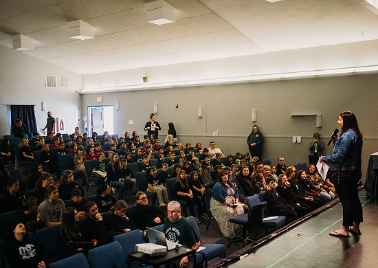 A speaker addresses a full auditorium of attentive students. The room is bright with overhead lighting, and the audience appears engaged and curious.