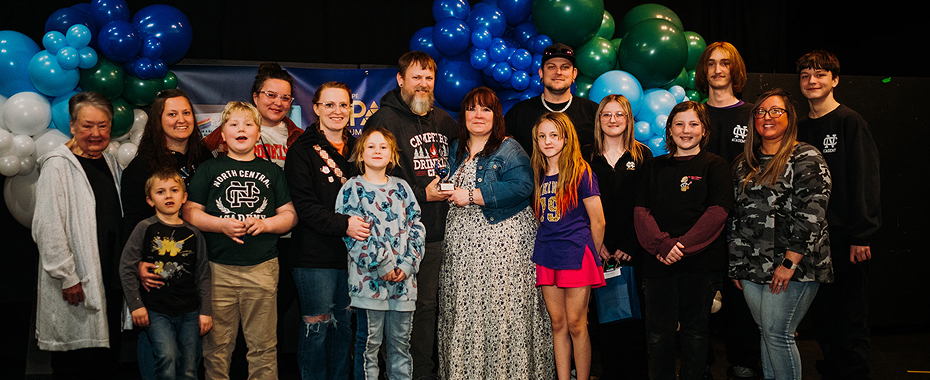 A diverse group of people of various ages stands in front of blue and white balloons, smiling for a group photo on a stage, conveying a celebratory mood.
