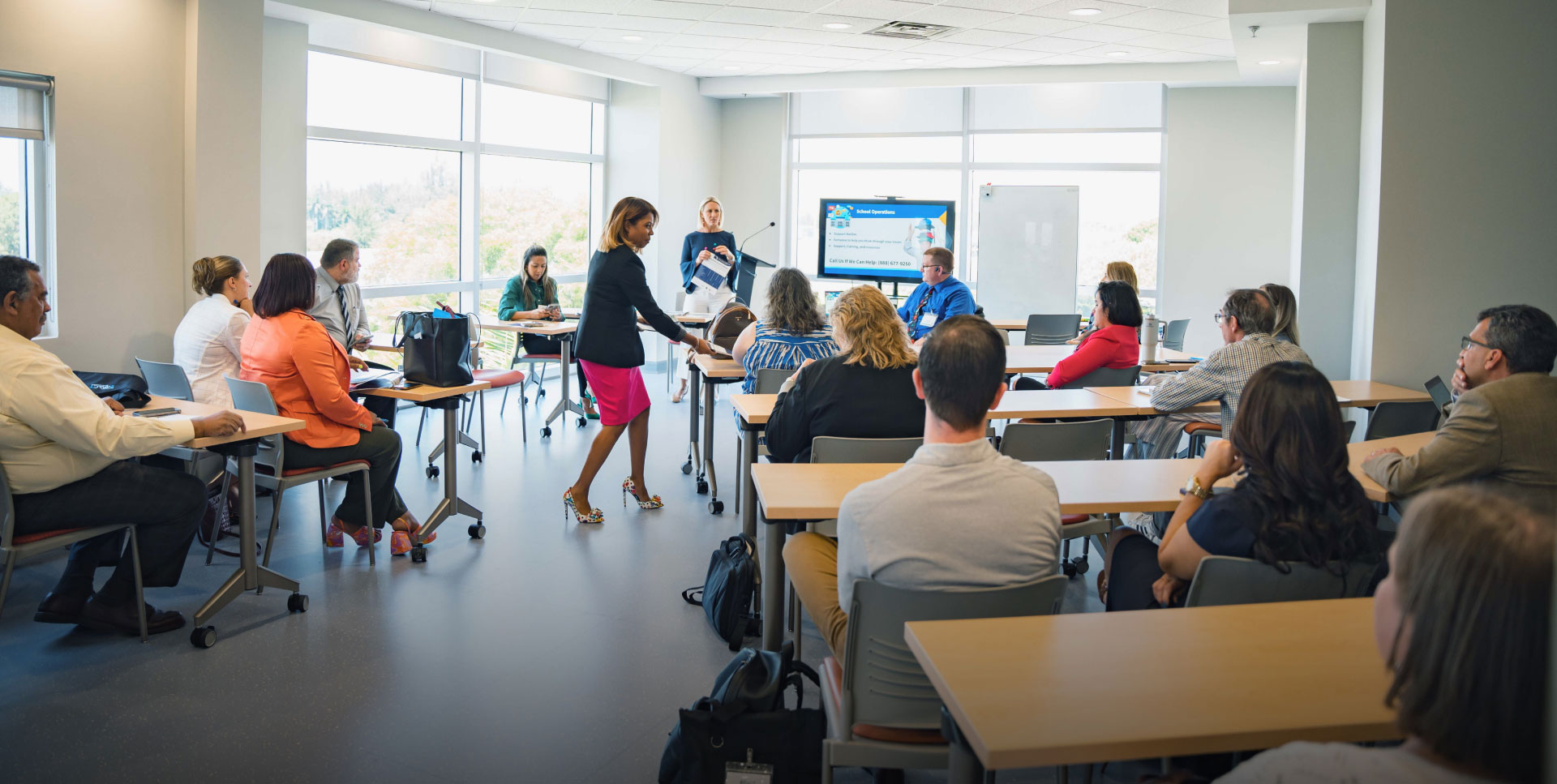 A group of people in a bright conference room listens attentively to a woman presenting. She stands by a screen displaying slides. The atmosphere is focused.