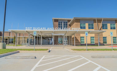 Modern school building with "SST Sugar Land College Prep" sign. Brick facade, glass entrance, clear blue sky, and accessible parking spaces in the foreground.