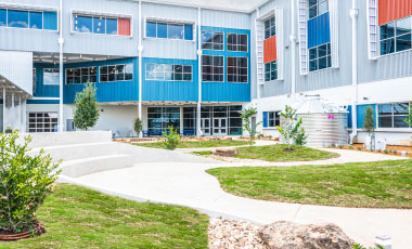 Modern school courtyard with blue, red, and gray building facade, featuring green lawns, small trees, and curving walkways under bright sunlight.