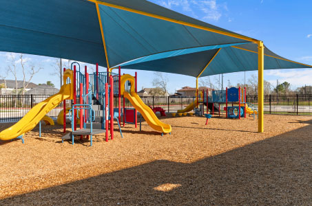 A colorful playground with red, yellow, and blue slides sits under large blue shade canopies. The ground is covered with wood chips, under a clear blue sky.