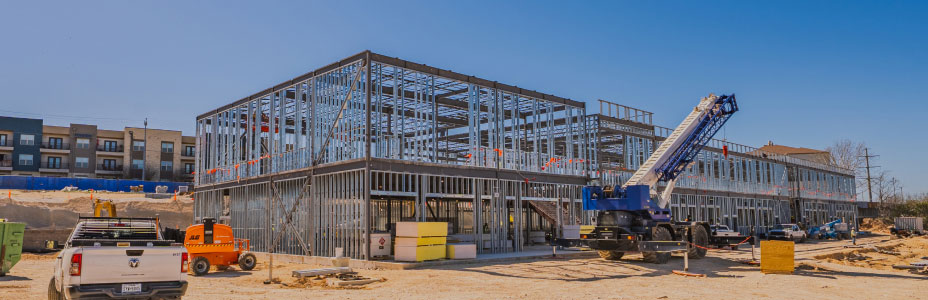 Construction site with a steel frame structure for a large building under a clear blue sky. A crane is positioned nearby, and a truck is parked in the foreground.