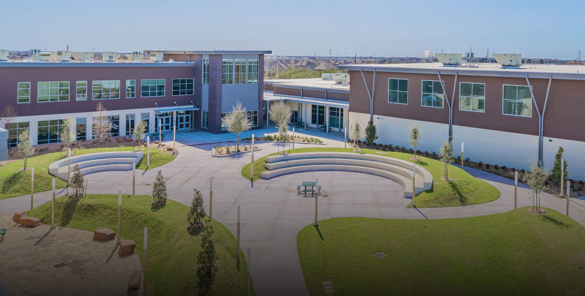 Modern school courtyard with circular seating, surrounded by green lawns. The two-story building has large windows, creating an inviting and airy atmosphere.