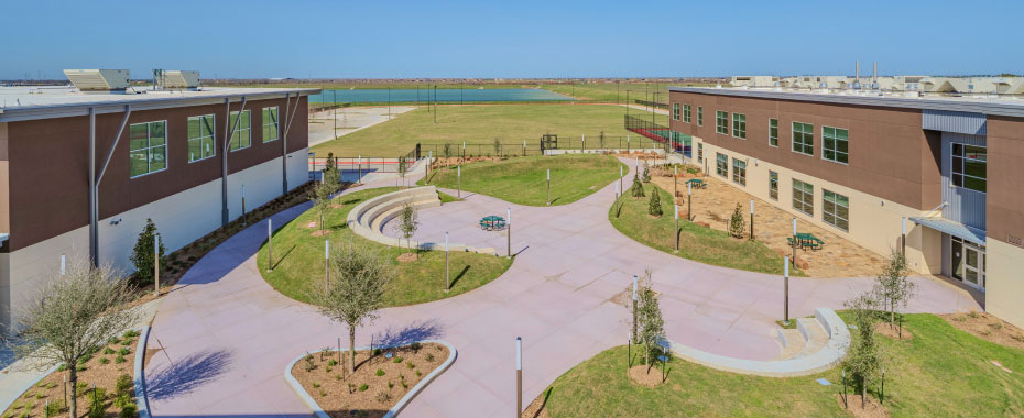 Aerial view of a modern school campus with two-story buildings, green lawns, circular walkways, and small amphitheater, under a clear blue sky.