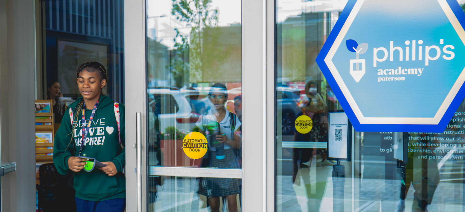 Students exit a school building, with one smiling girl in the foreground holding a green cup. A "Philip's Academy" sign is visible on the glass door.