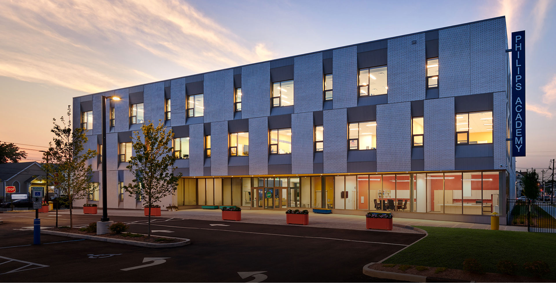 Three-story building labeled "Philips Academy" at dusk, with illuminated windows and a modern facade. Sparse trees and an empty parking lot in front.