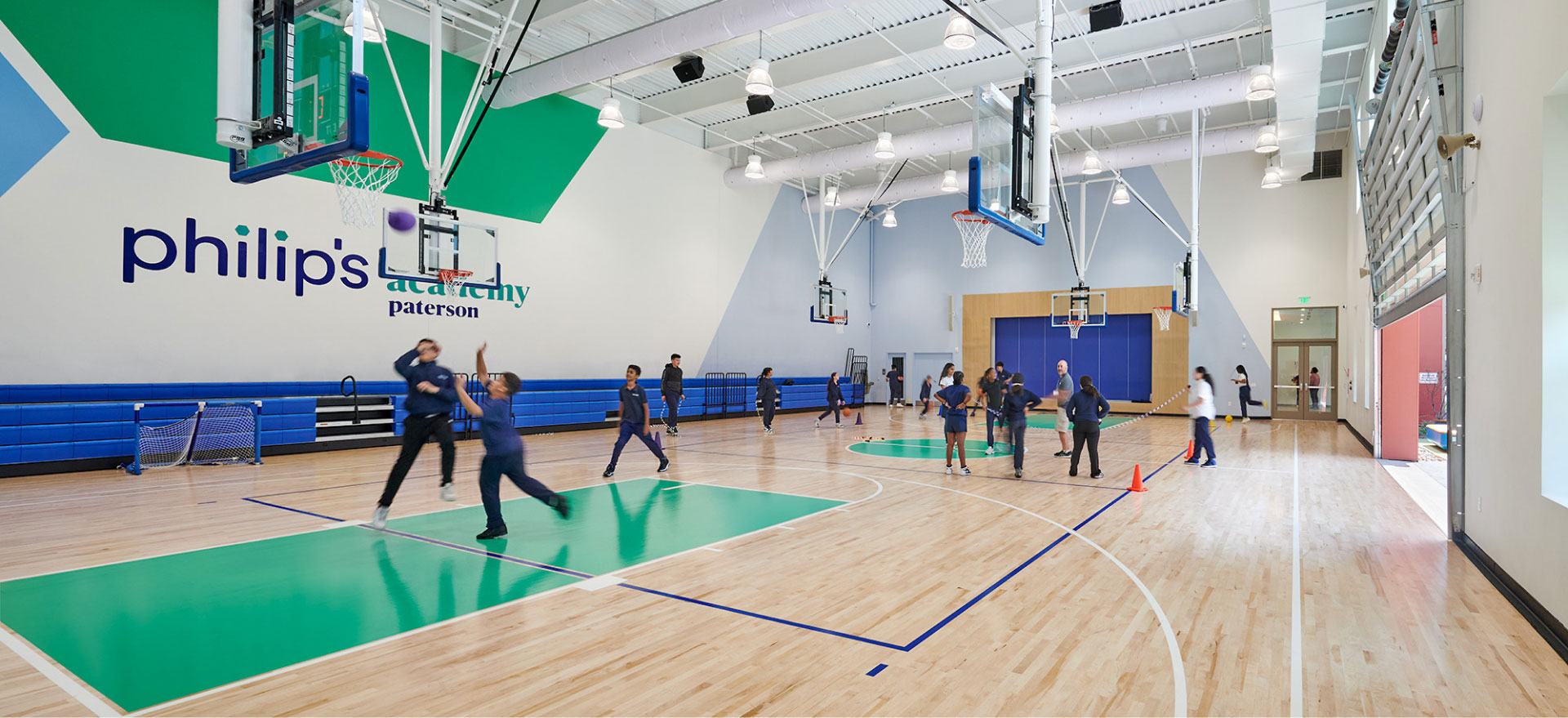 Students in gym uniforms play basketball in a bright gym with a wooden floor and green accents. The atmosphere is lively and active.