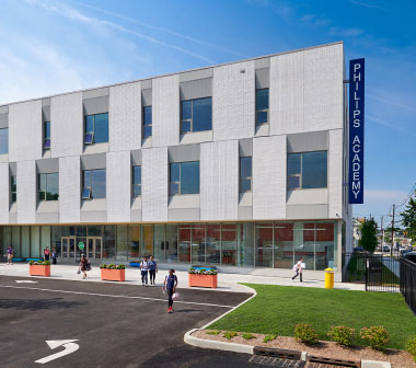 Modern three-story school building with geometric windows, "PHILLIPS ACADEMY" sign. Students walk outside on a sunny day, conveying an inviting atmosphere.