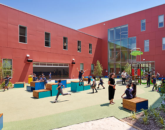 Outdoor school courtyard with red buildings. Children are playing on benches and a jungle gym. Adults supervise. Atmosphere is lively and sunny.