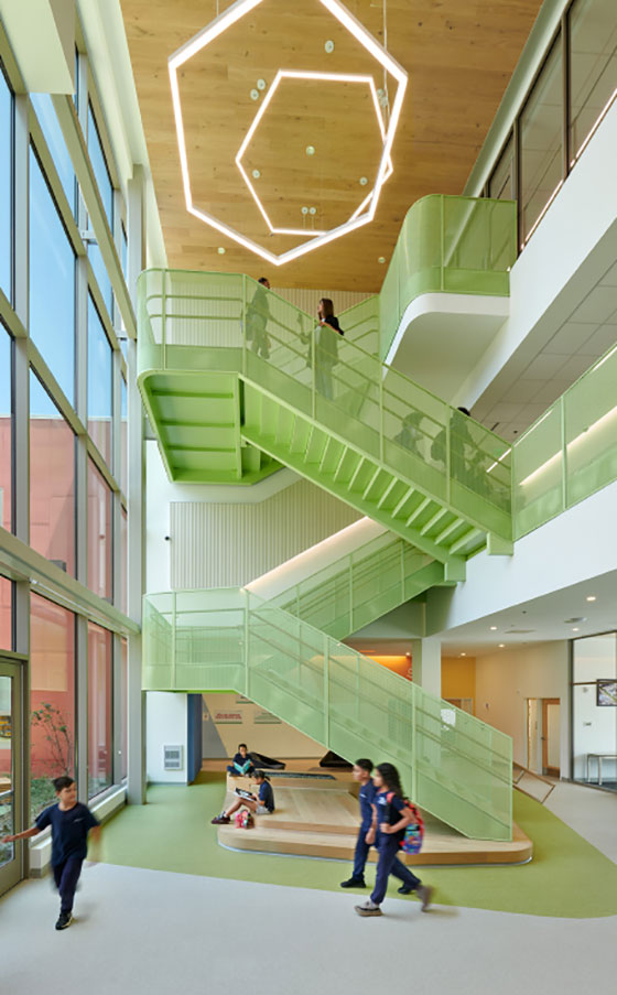 Bright, modern atrium with green geometric staircase, students walking, and a large, hexagonal light fixture above. The space feels open and vibrant.